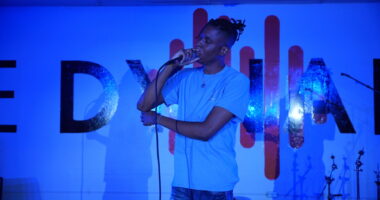 A man stands on stage singing into a microphone. The stage is lit with a blue light.