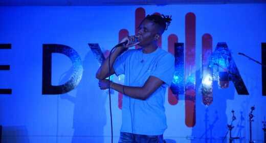 A man stands on stage singing into a microphone. The stage is lit with a blue light.