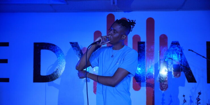 A man stands on stage singing into a microphone. The stage is lit with a blue light.