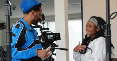 A man dressed in blue holds a video camera and talks with a woman who's smiling broadly and looks to be animated while talking. Given the camera gear visible, it's clear the two are doing a documentary film shoot.