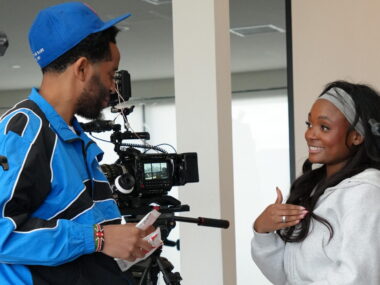A man dressed in blue holds a video camera and talks with a woman who's smiling broadly and looks to be animated while talking. Given the camera gear visible, it's clear the two are doing a documentary film shoot.