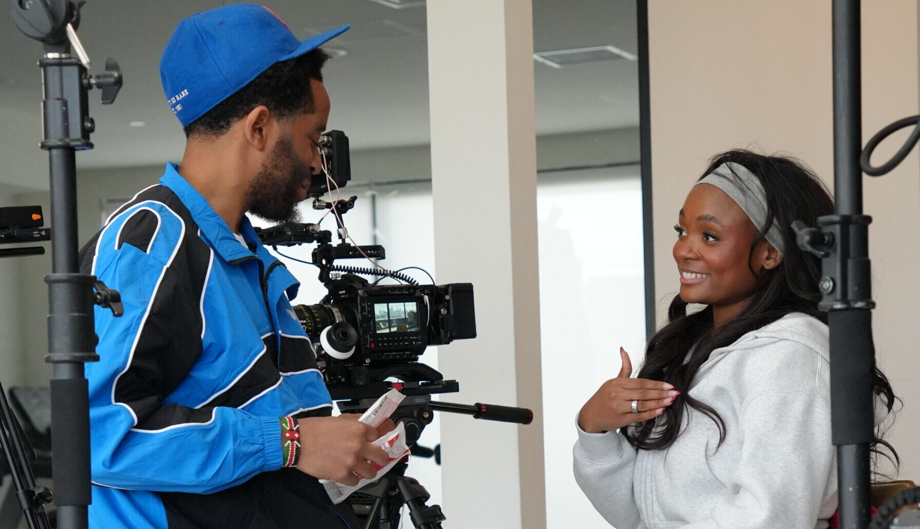 A man dressed in blue holds a video camera and talks with a woman who's smiling broadly and looks to be animated while talking. Given the camera gear visible, it's clear the two are doing a documentary film shoot.