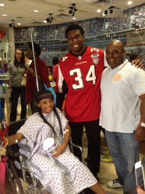 A young woman of high school age sits in a wheelchair wearing a hospital gown. Next to her are two smiling men, one of which is wearing a red football jersey with the number 34.