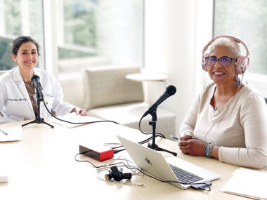 Two men pause from recording a podcast to smile at the camera. Both have microphones in front of them, and they are seated in a brightly lit (with natural sunlight) room with large windows. They're seated at a large table, and the woman on the left is wearing a white lab coat.
