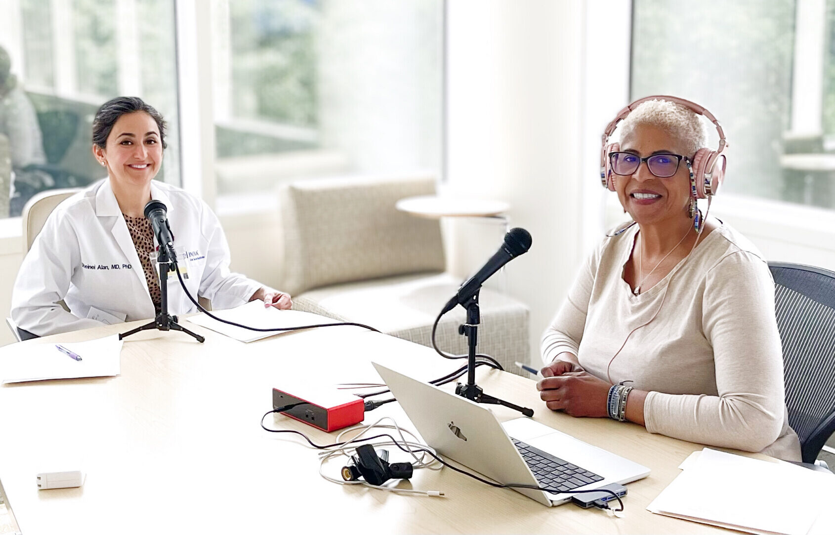 Two men pause from recording a podcast to smile at the camera. Both have microphones in front of them, and they are seated in a brightly lit (with natural sunlight) room with large windows. They're seated at a large table, and the woman on the left is wearing a white lab coat.