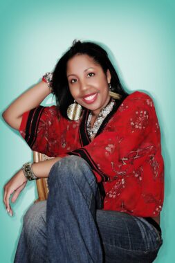 A playful portrait-style photograph of a woman sitting in a chair against a blue-green wall. Her pose is interesting, evoking curiosity, playfulness, and happiness. She has her right hand behind her head, and her left arm folded across her right knee; her right knee is crossed over her left knee. She's wearing blue jeans and a colorful blouse and accessories. 